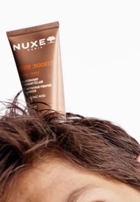Bruine tube van Nuxe Men [Boost] hydrator, met een textuur oppervlak en bedrukte branding, geplaatst op donker, rommelig haar.