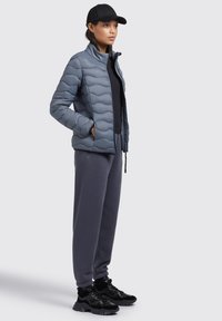 Grå puffjakke med bølgemønster, sort lynlås og sidelommer, parret med grå sweatpants og sorte sneakers, står oprejst.