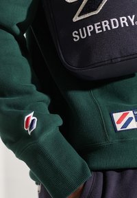 Groene trui met geribbelde manchetten, met een geborduurd logo in wit, rood en blauw op de mouw. Zwarte tas met duidelijk zichtbare tekst "SUPERDRY".