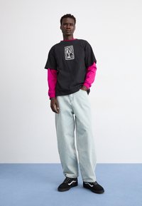 Černé oversized tričko s bílou grafikou, položené na růžovém tričku s dlouhým rukávem. Světle modré volné džíny a černé tenisky dotvářejí celkový vzhled.