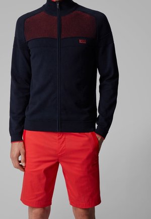 Mies, jolla on päällään tummansininen vetoketjullinen paita, jossa on teksturoidut punaiset paneelit, ja kirkkaanpunaiset polvipituiset shortsit, yksinkertaisen harmaan taustan edessä.