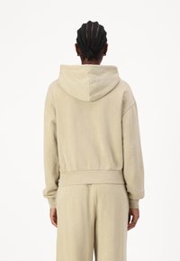 Beige hoodie med avslappnad passform, huva och ribbad mudd. Tyget verkar mjukt med minimal detaljer, vilket visar en enkel, mångsidig design.