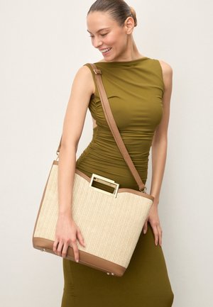Woman in olive green sleeveless dress carrying a large beige and tan woven tote bag with a shoulder strap, smiling and looking down.