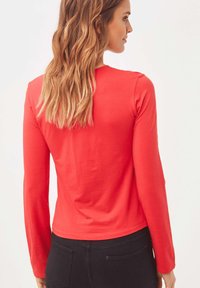 Camiseta roja de manga larga, hecha de algodón, presenta un diseño ajustado con un escote redondo y una textura suave, combinada con pantalones negros.