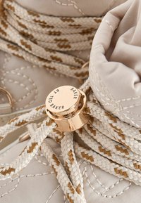 Béžová látková bota se zlatými akcenty, s opletenými tkaničkami a kulatou kovovou visačkou s gravírovaným nápisem "Ted Baker". Viditelný texturovaný šev.