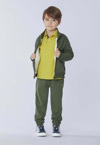 Giacca verde con zip e fodera in pile bianco, polo gialla, pantaloni della tuta verdi e sneaker bianche con dettagli blu.