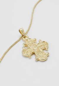 Złota zawieszka w kształcie etiopskiego krzyża z misternymi grawerunkami na delikatnym złotym łańcuszku, przedstawiona na jednolitym białym tle.