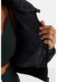 Mano con anillos de oro sosteniendo una chaqueta negra con cremallera abierta que muestra un forro interior de marca sobre una parte superior y pantalones de canalé de color verde oscuro.