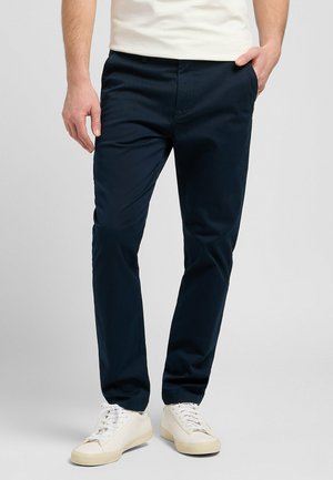 Navy blue chinos made of smooth fabric with a slim fit, featuring side pockets and a buttoned closure. Worn with white sneakers.