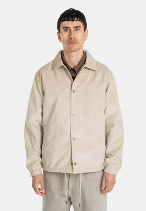 Mann mit kurzen dunklen Haaren trägt eine beige Jacke mit Knöpfen und passende Hosen, steht vor einem einfarbigen hellgrauen Hintergrund.