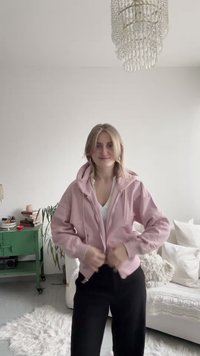 Hoodie zippé rose clair avec poche kangourou, cordons ajustables et manches longues, associé à un pantalon noir sur un tapis blanc texturé.