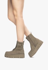 Bottines vert olive avec un dessus en daim texturé, un système de laçage à l'avant, une fermeture éclair sur le côté et une semelle épaisse à plate-forme avec des rainures.