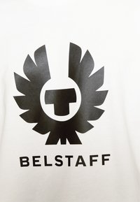 Balta t-krekls ar lielu melnu logotipu, kas attēlo emblēmu, kas atgādina spārnus, centrā virs teksta "BELSTAFF" melnā bieza fontā.