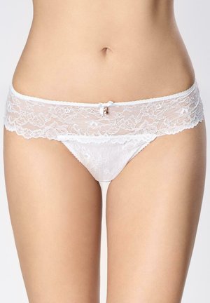 Kobieta w białych koronkowych majtkach z kwiatowym wzorem, niskim stanem oraz z małą kokardką i ozdobą na środku przodu.