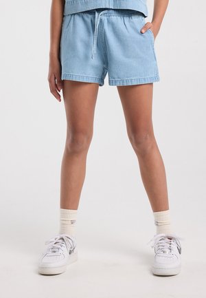 Person trägt hellblaue Jeansshorts, weiße Sneaker und cremefarbene Socken und steht mit einer Hand in der Tasche.