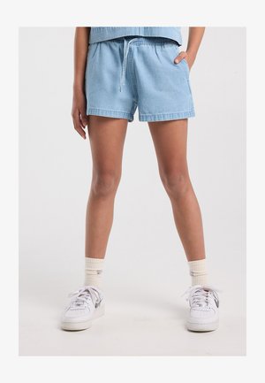 Persona con pantalones cortos vaqueros azul claro, zapatillas blancas y calcetines color crema, de pie con una mano en el bolsillo.