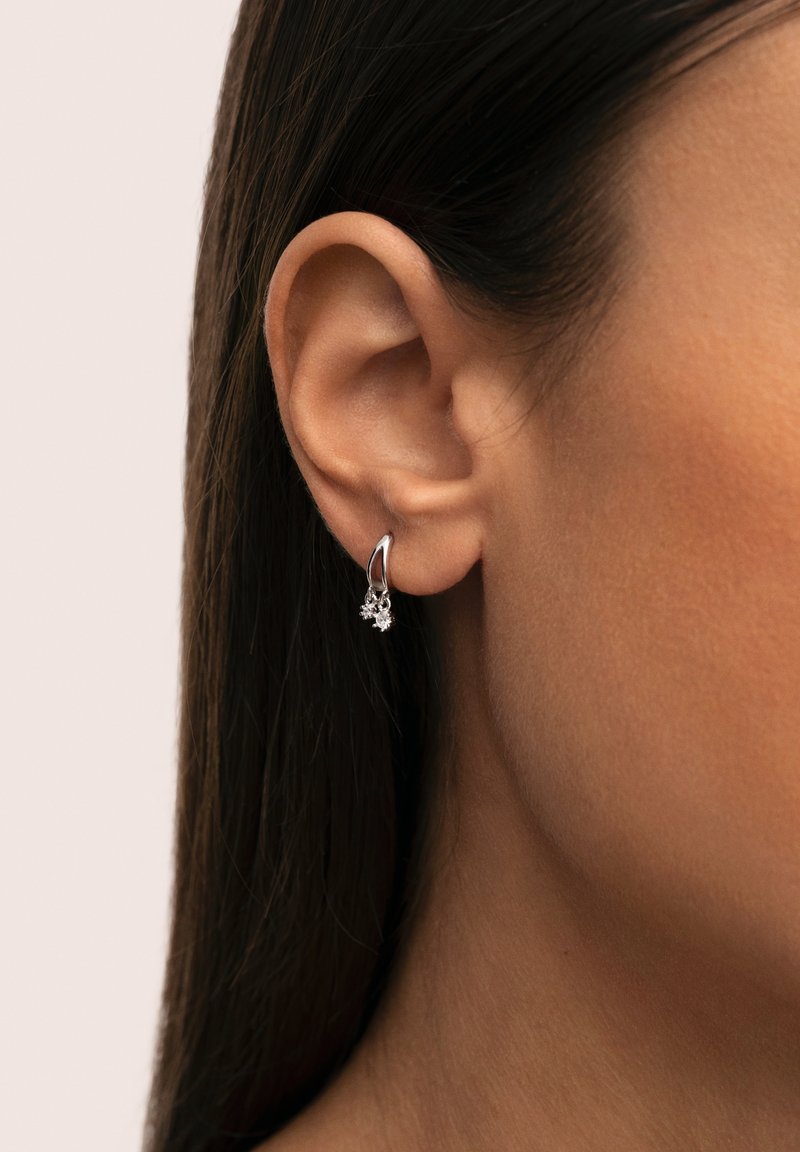 Aro de plata con un pequeño diseño floral y acentos brillantes, colgando de la oreja sobre un fondo neutro.
