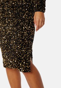 Černé šaty se třpytkami s přiléhavým střihem, zdobené zlatými třpytkami po celém povrchu a bočním rozparkem. Dlouhé rukávy a jemná textura.