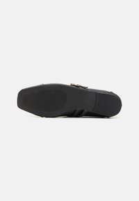 Schwarze Schuhsohle aus strukturiertem Gummi für Grip, mit einem glatten Obermaterial und dekorativen Schnallenakzenten an der Seite.