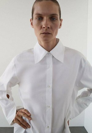 Camicia bianca con colletto classico, maniche lunghe con spacchi laterali e una texture del tessuto liscia. Design semplice e privo di decorazioni.