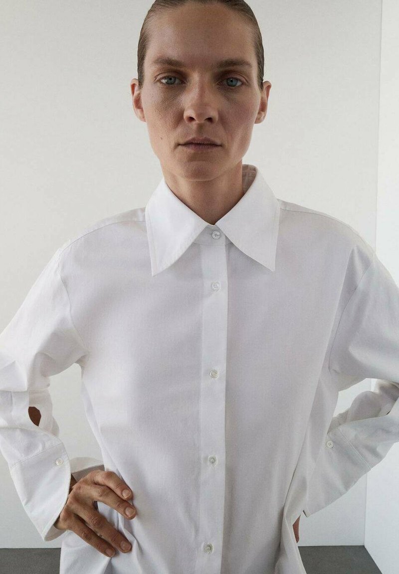 Camisa blanca de botones con cuello clásico, mangas largas que presentan aberturas laterales y una textura de tela suave. Diseño simple y sin adornos.