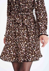 Robe fleurie marron avec de petites fleurs roses, blanches et bleues, taille cintrée avec une ceinture assortie, et manches à volants avec bordure en dentelle.