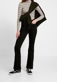 Höga svarta utsvängda jeans med vita sömmar, matchade med en randig långärmad topp och en olivgrön stickad tröja hängande över axlarna.