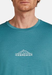 Mann in einem türkisfarbenen Rundhalsausschnitt-T-Shirt mit weißem "Icebreaker"-Text und Berglogo auf der Brust, ausgeschnitten auf Höhe des Halses und des oberen Oberkörpers.