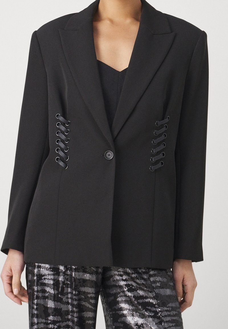 Blazer noir à un bouton avec de larges revers et des détails lacés de chaque côté, porté avec un pantalon noir à motif pailleté.