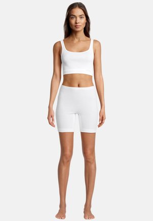 Frau steht barfuß und trägt ein weißes, kurzes Tanktop sowie weiße, enganliegende Shorts vor einem schlichten, hellen Hintergrund.