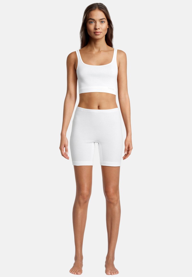 Kvinde stående barfodet iført en hvid kort tanktop og hvide tætsiddende shorts mod en ensartet lys baggrund.