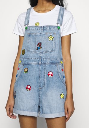 Krótkie ogrodniczki z denimu z naszywkami tematycznymi Super Mario, noszone na białej koszulce na osobie z rozluźnionymi ramionami wzdłuż ciała.