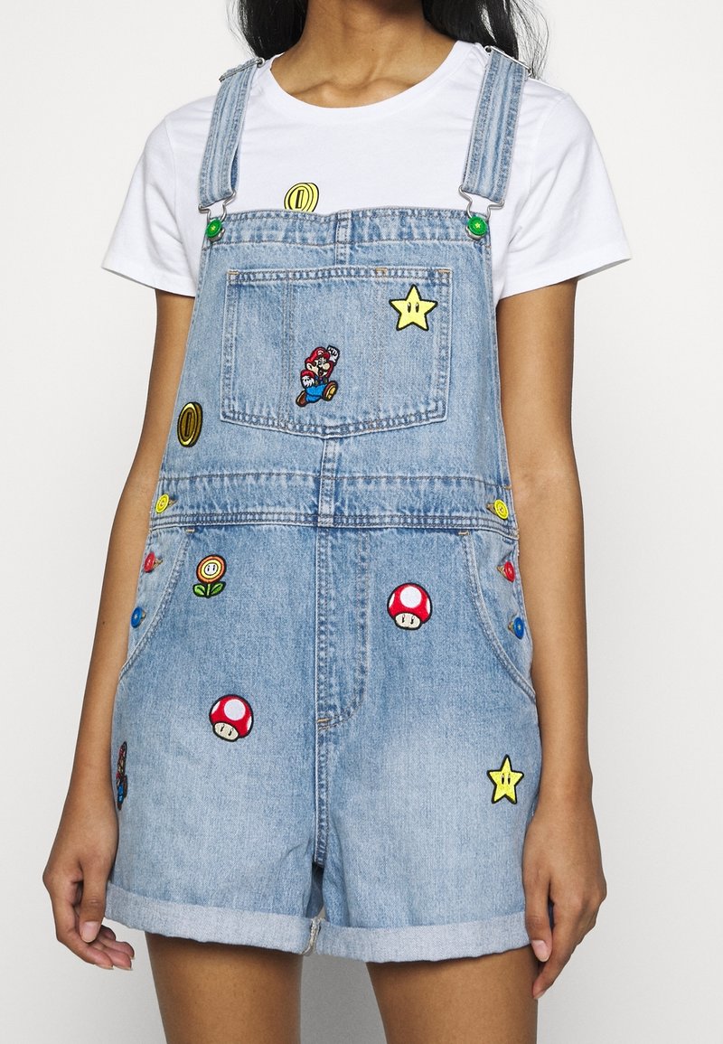 Salopette courte en denim avec des patchs sur le thème de Super Mario portée sur un t-shirt blanc par une personne aux bras détendus le long du corps.