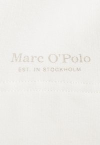 Tela blanca con una textura suave que presenta el logo "Marc O'Polo" y "EST. IN STOCKHOLM" impreso en gris claro.