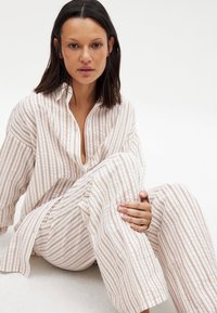 Luźny, pasiasty komplet piżamy w miękkim beżowym i białym materiale. Zapięcie na guziki przy kołnierzyku oraz szerokie rękawy z pionowymi liniami.