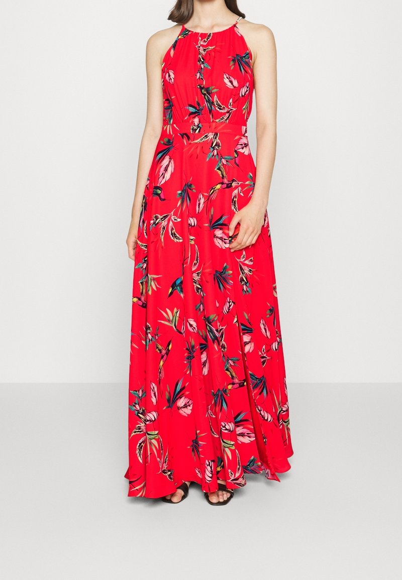Femme portant une robe maxi rouge sans manches avec un motif floral tropical, debout devant un fond clair uni.