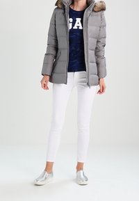 Grijze pufferjas met een capuchon van nepbont, gecombineerd met een marineblauw shirt met witte tekst, witte strakke jeans en zilveren instapschoenen.