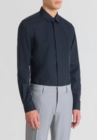 Camicia a maniche lunghe blu navy con colletto button-down; realizzata in tessuto liscio, con un design aderente e una leggera lucentezza. Indossata con pantaloni grigi.