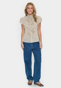 Blusa beige con volant, maniche corte e colletto alla coreana, abbinata a jeans in denim blu larghi e scarpe piumate con motivo.