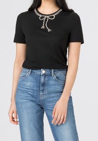 Svart kortärmad t-shirt med en dekorativ krage av rhinstenar och rosettdetalj, kombinerad med högmidjade blå denimjeans.