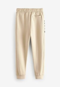 Pantalones de chándal beige con cintura elástica, puños acanalados, bolsillo lateral y detalle de logo a lo largo del lado izquierdo. Textura de tejido suave y liso.