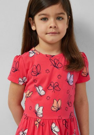 Jeune fille aux longs cheveux bruns portant une robe rouge avec des motifs de papillons multicolores, debout devant un fond uni.