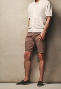 Camisa de punto blanca de manga corta con un patrón texturizado. Pantalones cortos marrones con puños enrollados. Zapatos negros sin cordones con detalles trenzados.