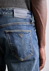 Kék farmernadrág, hátul zsebbel, amelyen gravírozott Emporio Armani logó és narancssárga varrás található, látható bőr márkajelzés a deréknál.