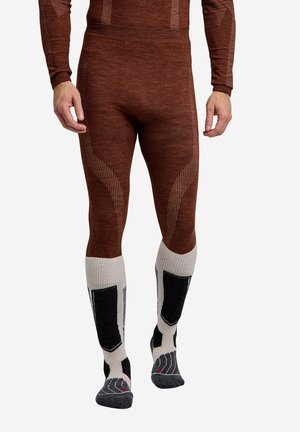 Man met bruine thermoleggings en lange sokken in wit en zwart, staand tegen een witte achtergrond.