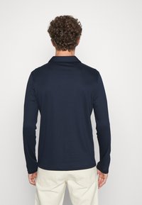 Marineblå langermet polo med krage, laget av glatt stoff. Bakvinduet viser en skreddersydd passform og subtile skul seams.