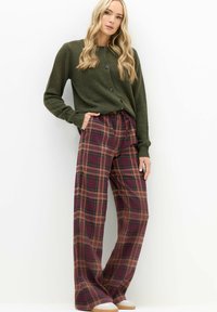 Oliivinvihreä neulecardigan, jossa on nappikiinnitys, yhdistettynä laajahelmaisiin ruudullisiin housuihin, joissa on burgundin, ruskean ja vihreän sävyjä. Valkoiset tennarit.