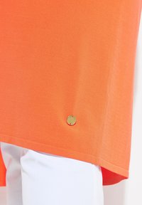 Orange bomullströja med en något böjd fåll och en gyllene logotyp detaj near den nedre vänstra hörnet. Bärs över vit tyg.