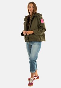 Giovane donna che indossa una giacca verde oliva con una toppa rosa, jeans blu arrotolati e sneaker bordeaux su uno sfondo bianco.