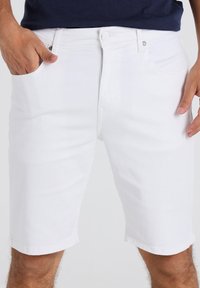 Hombre que lleva pantalones cortos de mezclilla blancos con las manos cerca de los bolsillos frontales, acompañado de una camiseta azul oscura, con las piernas visibles hasta la mitad del muslo.